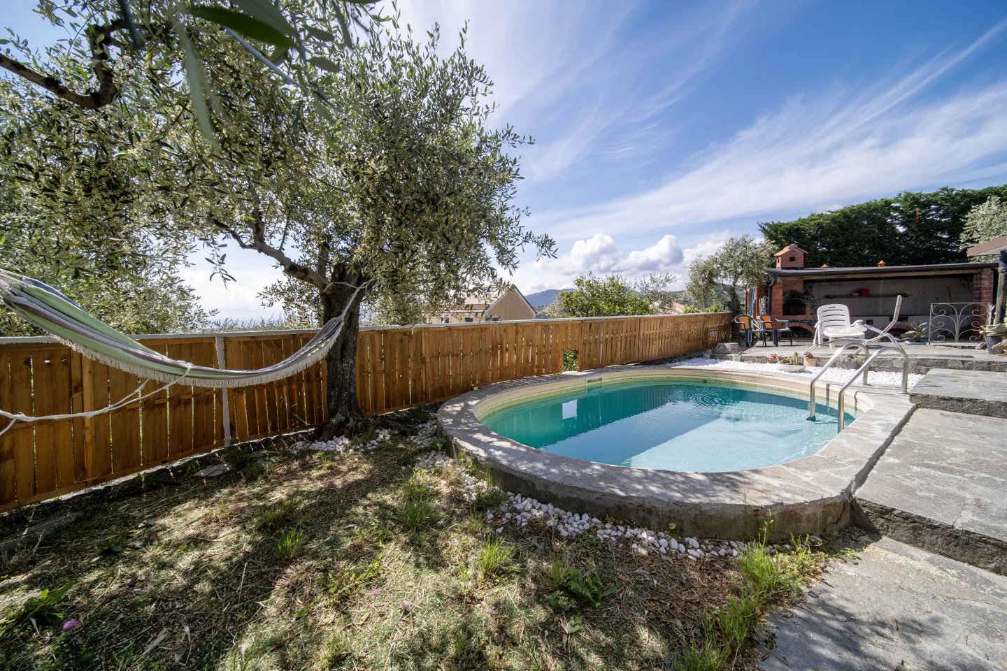 Backyard with a small pool, a hammock hanging from a tree, and a wooden fence under a blue sky.