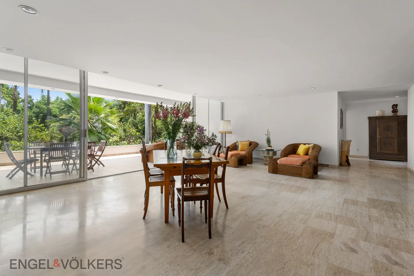 Bright, open living space with marble floors, dining table, wicker chairs, and a large sliding glass door to a patio with outdoor seating.