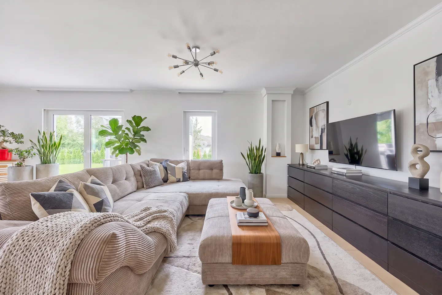 Bright living room with a large, beige sectional sofa, patterned pillows, and a matching ottoman. A black media console sits beneath a TV and artwork.