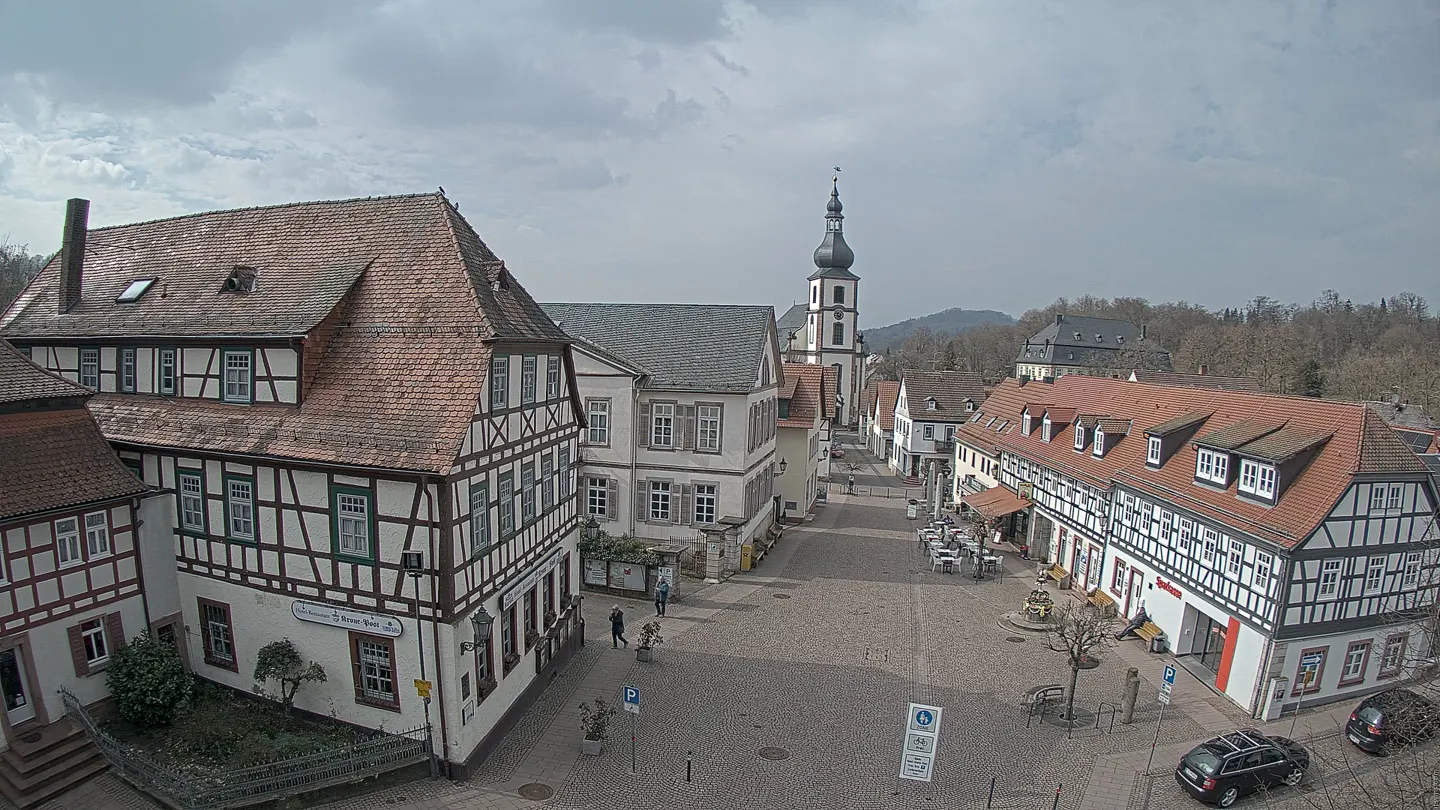 Town square with half-timbered buildings, a church steeple, and parked cars on a cloudy day.