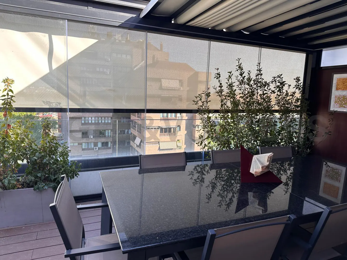 Outdoor dining area with a black table, gray chairs, and plants. Buildings are visible through the window shades.