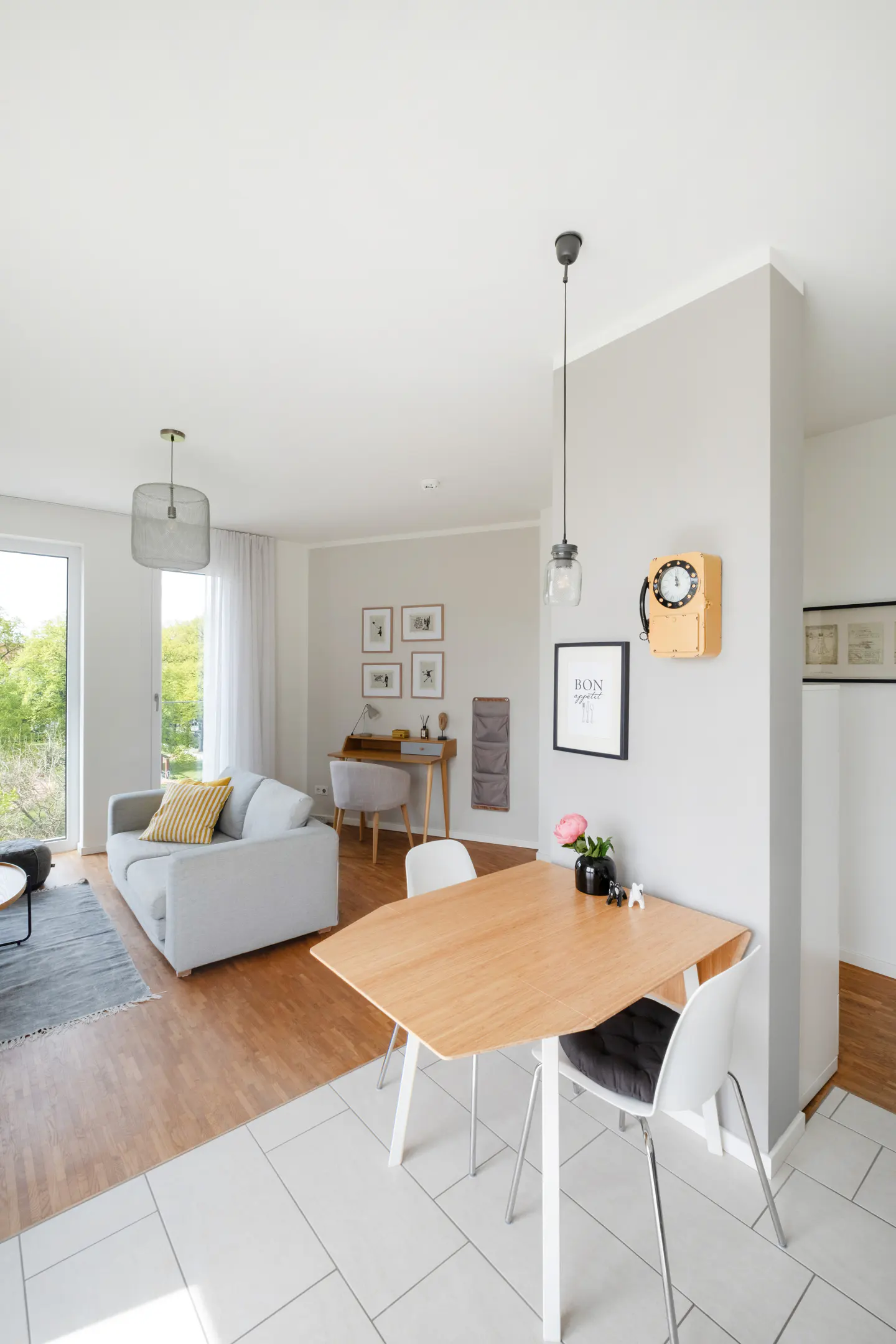 Bright, open-concept living space with a light wood dining table and white chairs in the foreground. A gray sofa and desk are visible in the background.