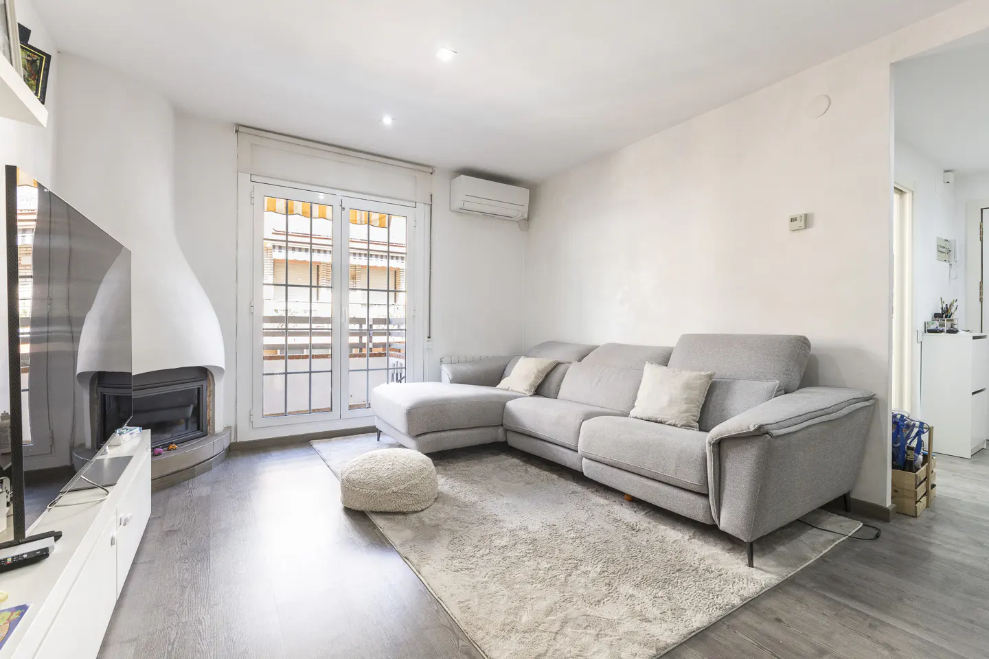 Bright living room with gray sectional sofa, white walls, fireplace, TV, and balcony doors. A light rug covers the gray wood floor.