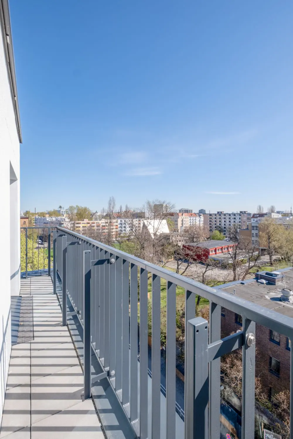 Balcony view with gray metal railing, light gray tiled floor, and city skyline under a clear blue sky.