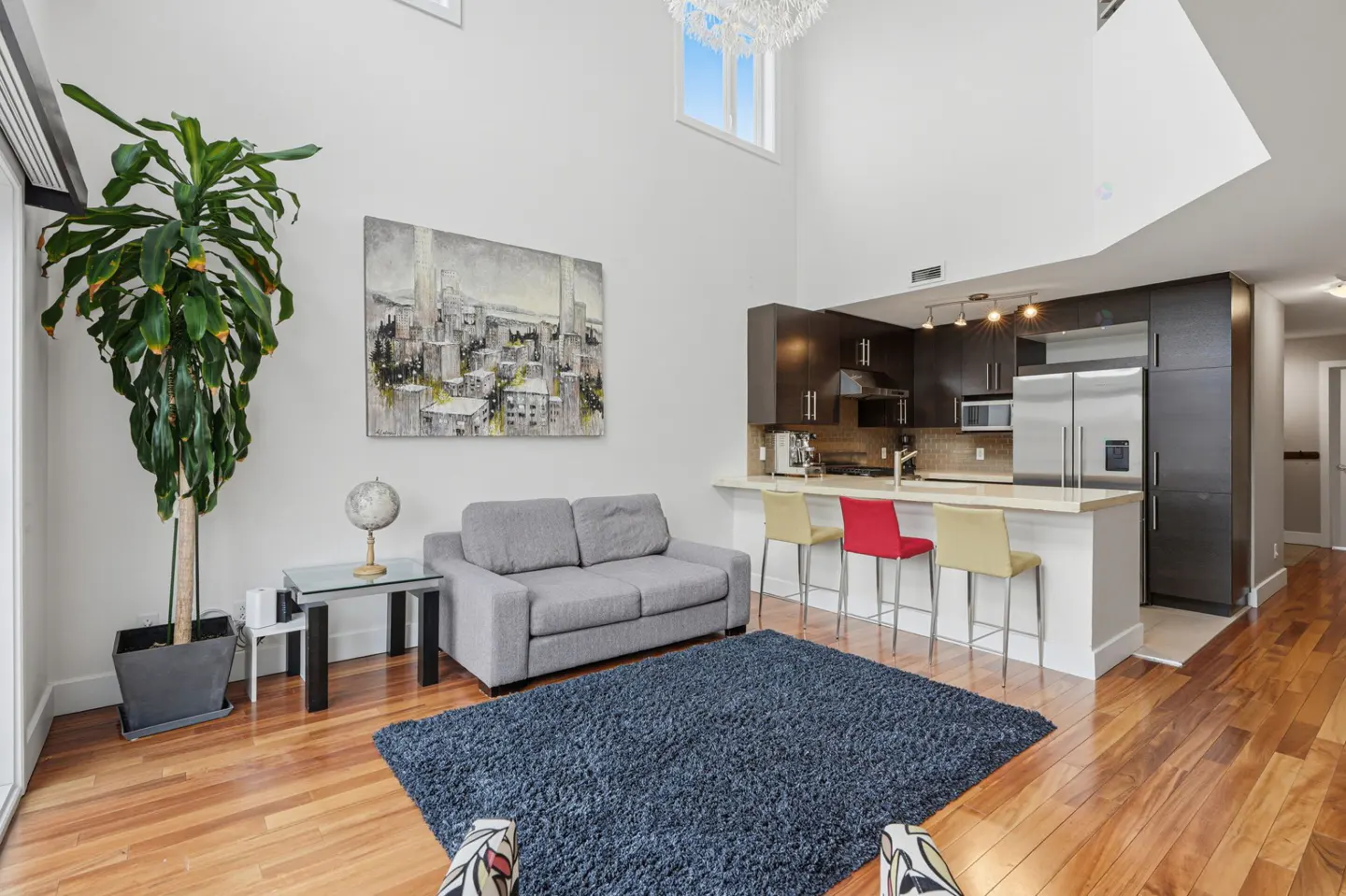 Bright, modern apartment interior with hardwood floors, a gray sofa, and a dark blue rug. A kitchen with bar seating is in the background.