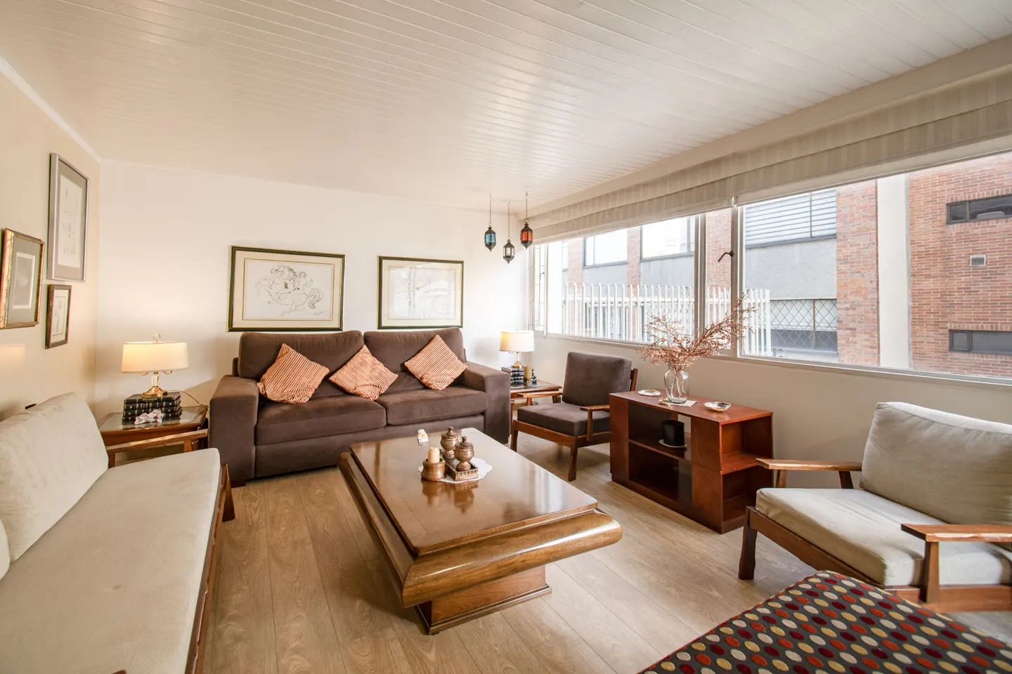 Bright living room with brown sofa, wood floors, and large windows. Art hangs on the walls, and a wooden coffee table sits in the center.