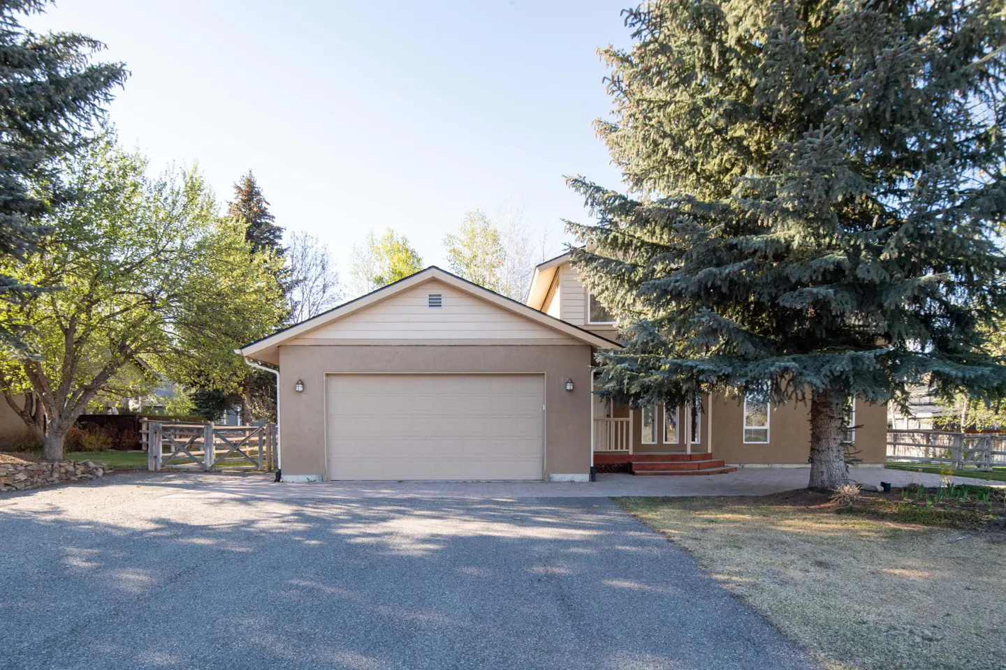 Beige house with a two-car garage and a paved driveway, surrounded by green trees.