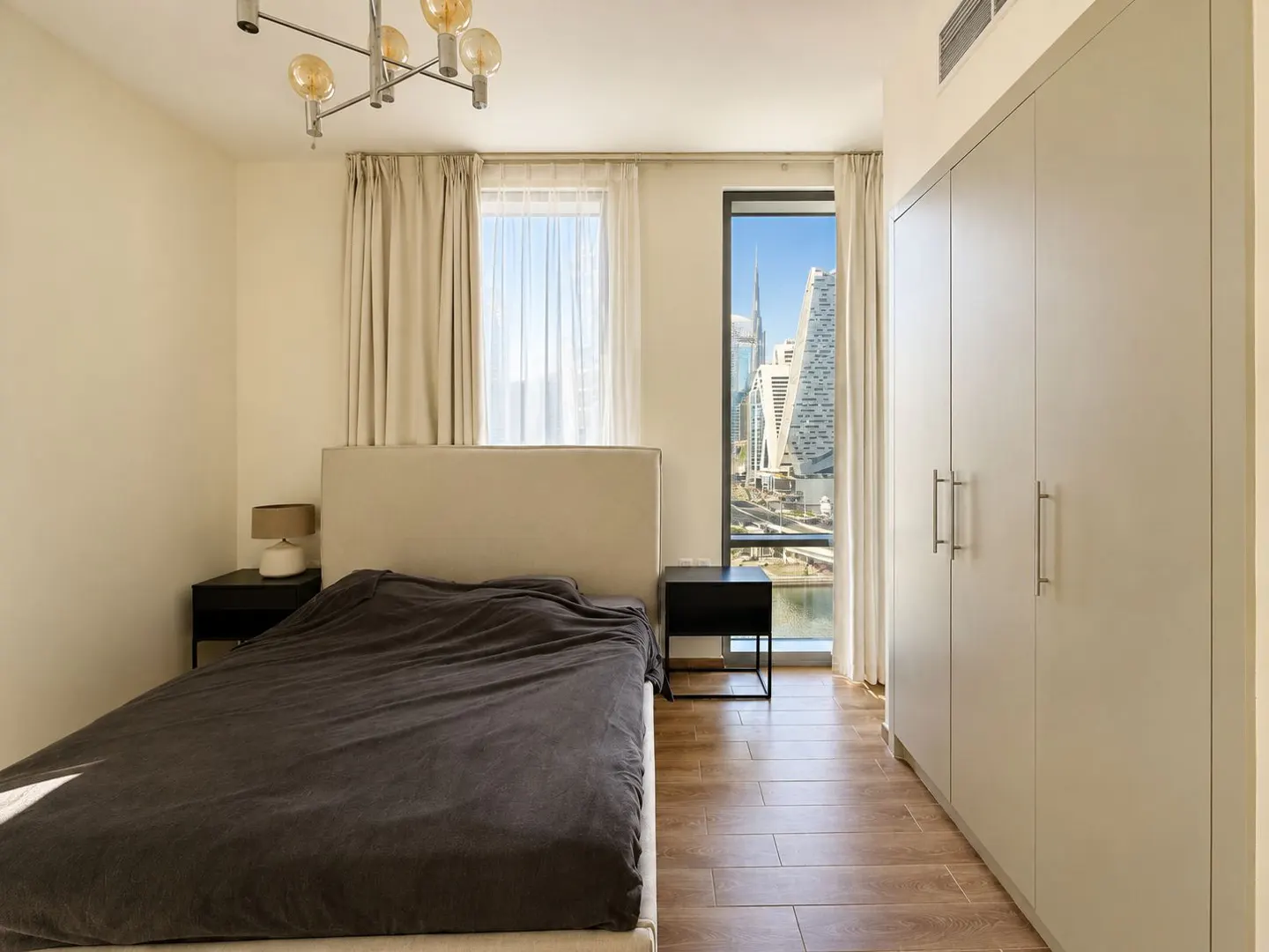 Bedroom with a bed, nightstands, and a view of Dubai's skyline through a window. The room has light walls and wood floors.