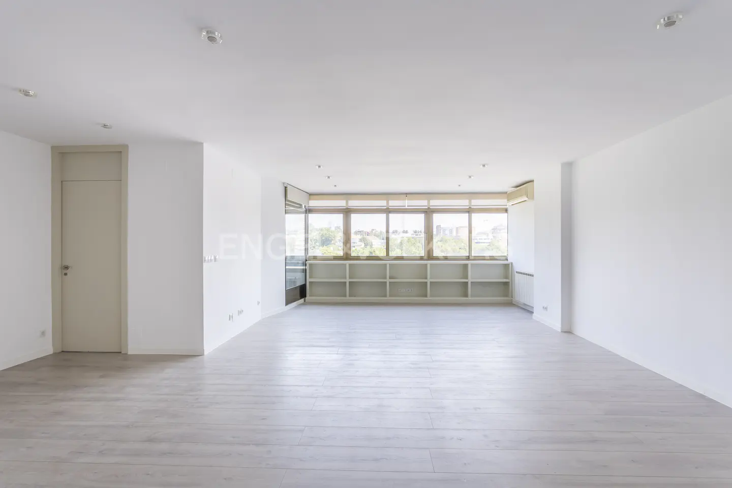 Bright, empty room with light wood floors, white walls, and a wall of windows with built-in shelving. A door is visible on the left.