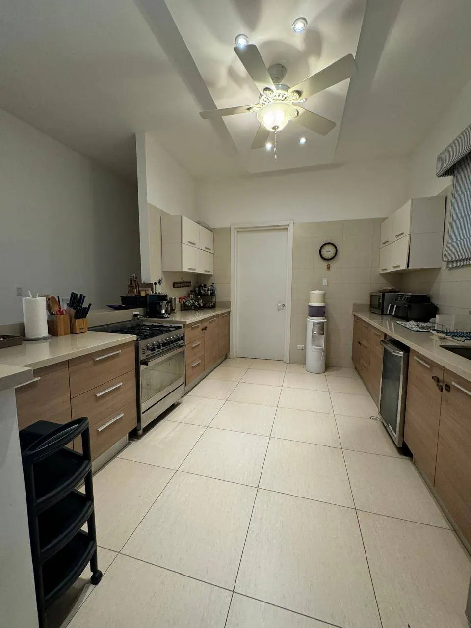 A spacious kitchen with light brown cabinets, stainless steel appliances, and beige tile flooring. A ceiling fan hangs above.
