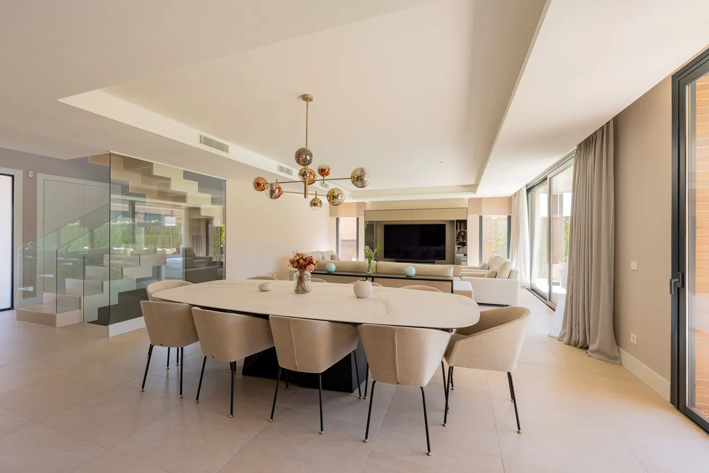 Open-concept living space with a white oval dining table, beige chairs, and a modern chandelier. A glass-railed staircase is visible.