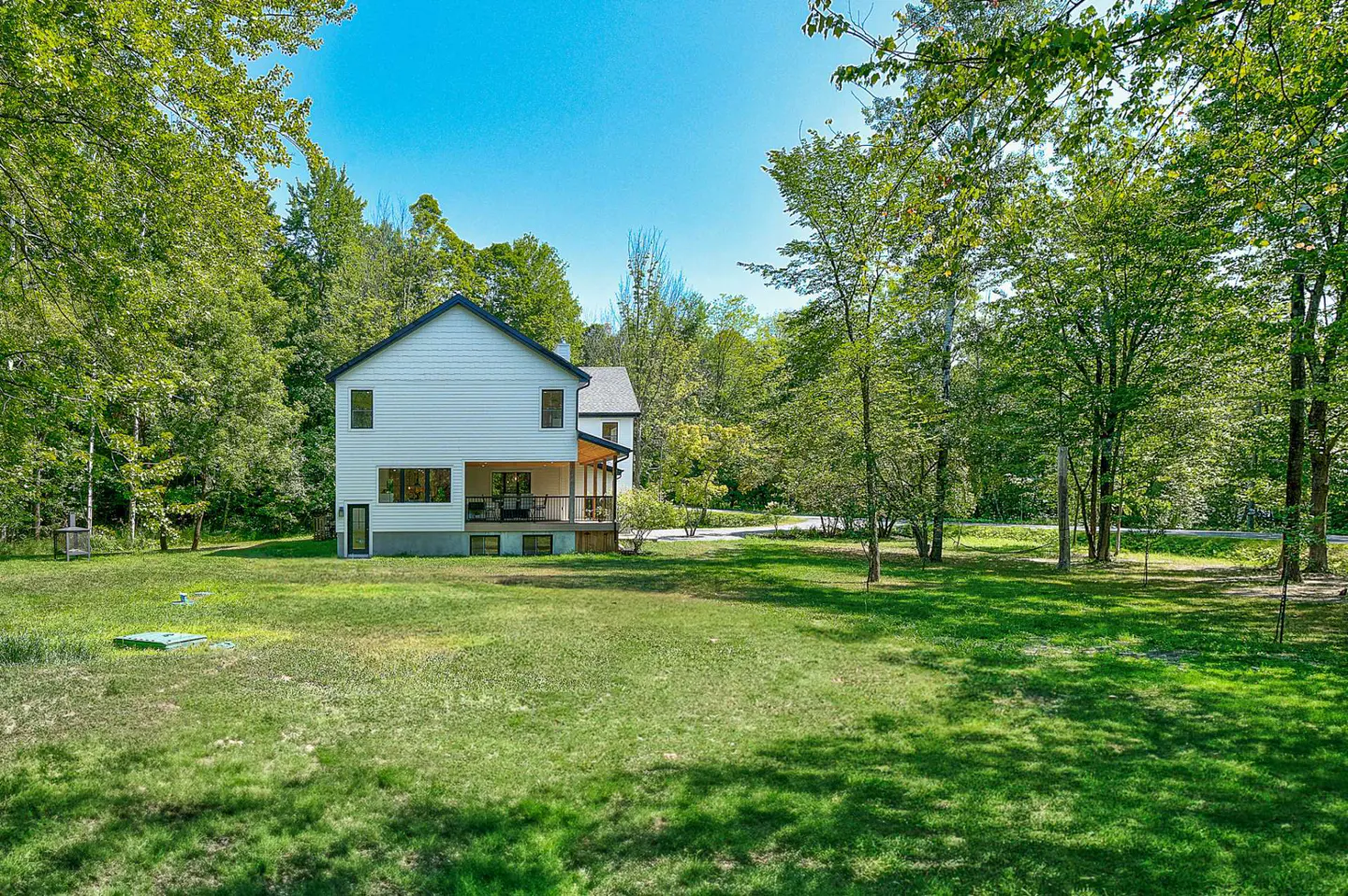 White two-story house with a dark roof and a covered porch, surrounded by green grass and trees under a blue sky.