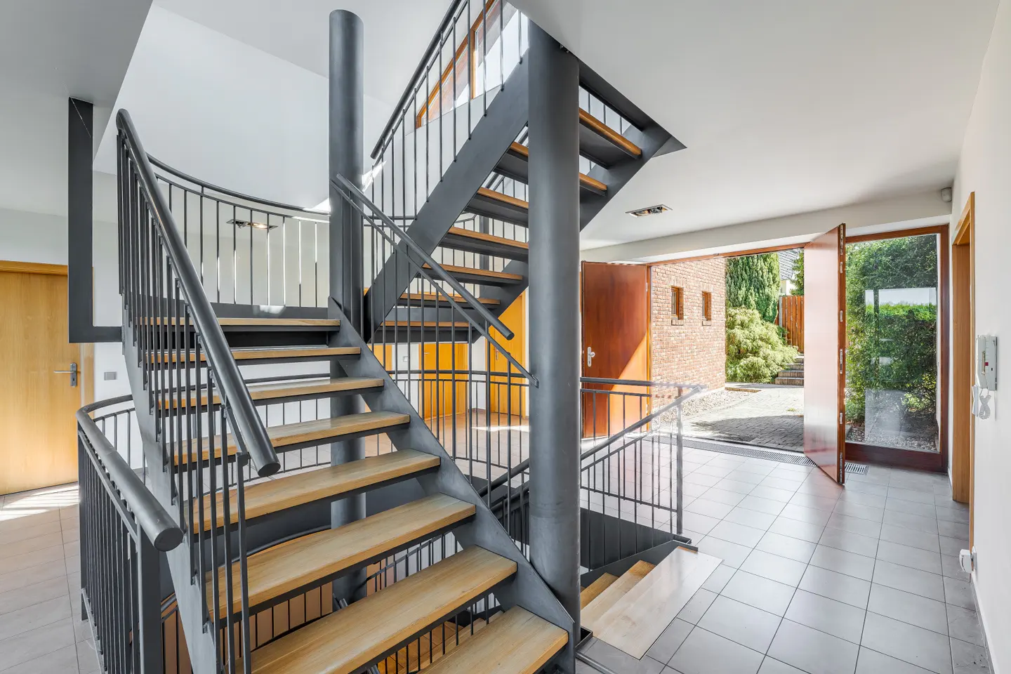 Interior view of a modern home featuring a metal staircase with wooden steps, leading to an open doorway with a view of a garden.