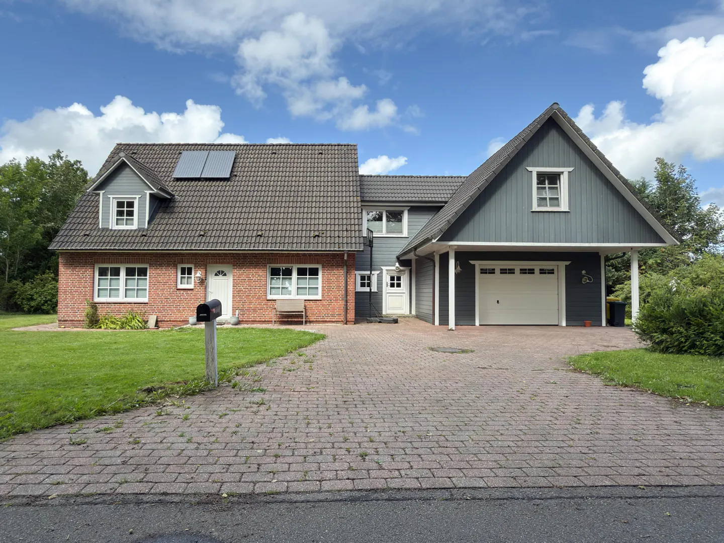 Two-story home with brick and gray siding, a brick driveway, and a green lawn under a blue sky with white clouds.