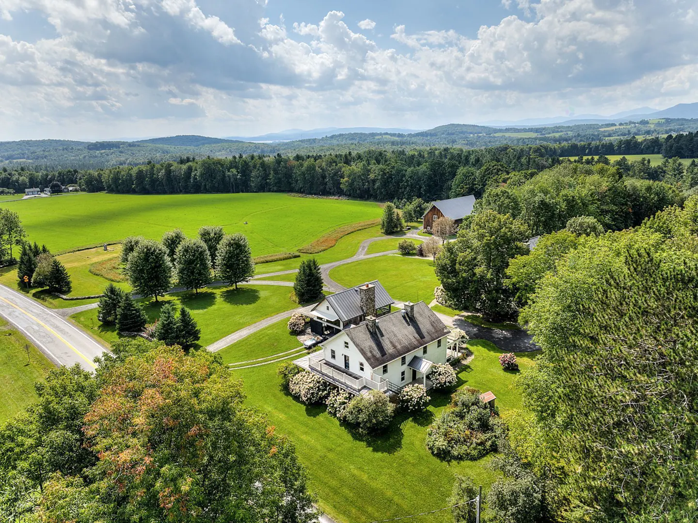 Aerial view of a white farmhouse with a gray roof, surrounded by green lawns, trees, and a large green field. Mountains are visible in the distance.
