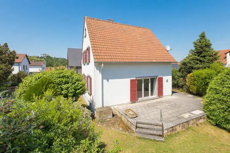 White house with a red-tiled roof and red shutters, featuring a stone patio and steps in a sunny, green residential area.
