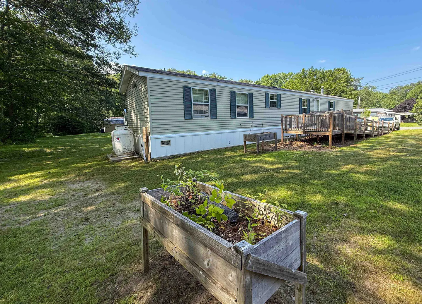 A single-story mobile home with light green siding and blue shutters, a wooden deck, and a raised garden bed in the yard.