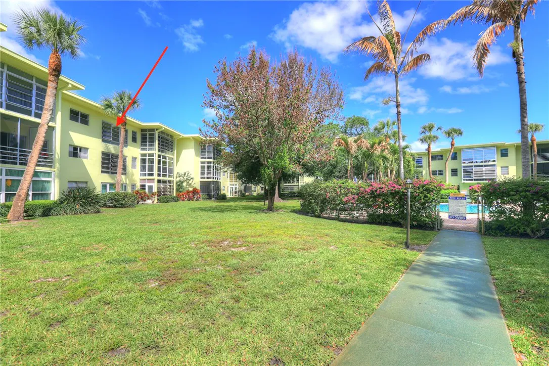 Exterior view of a light yellow apartment complex with palm trees and a green lawn under a blue sky.