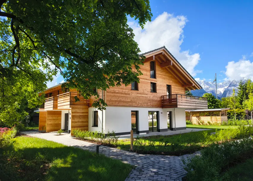 Two-story house with wood siding and white base, balcony, green lawn, and mountain view under a blue sky.