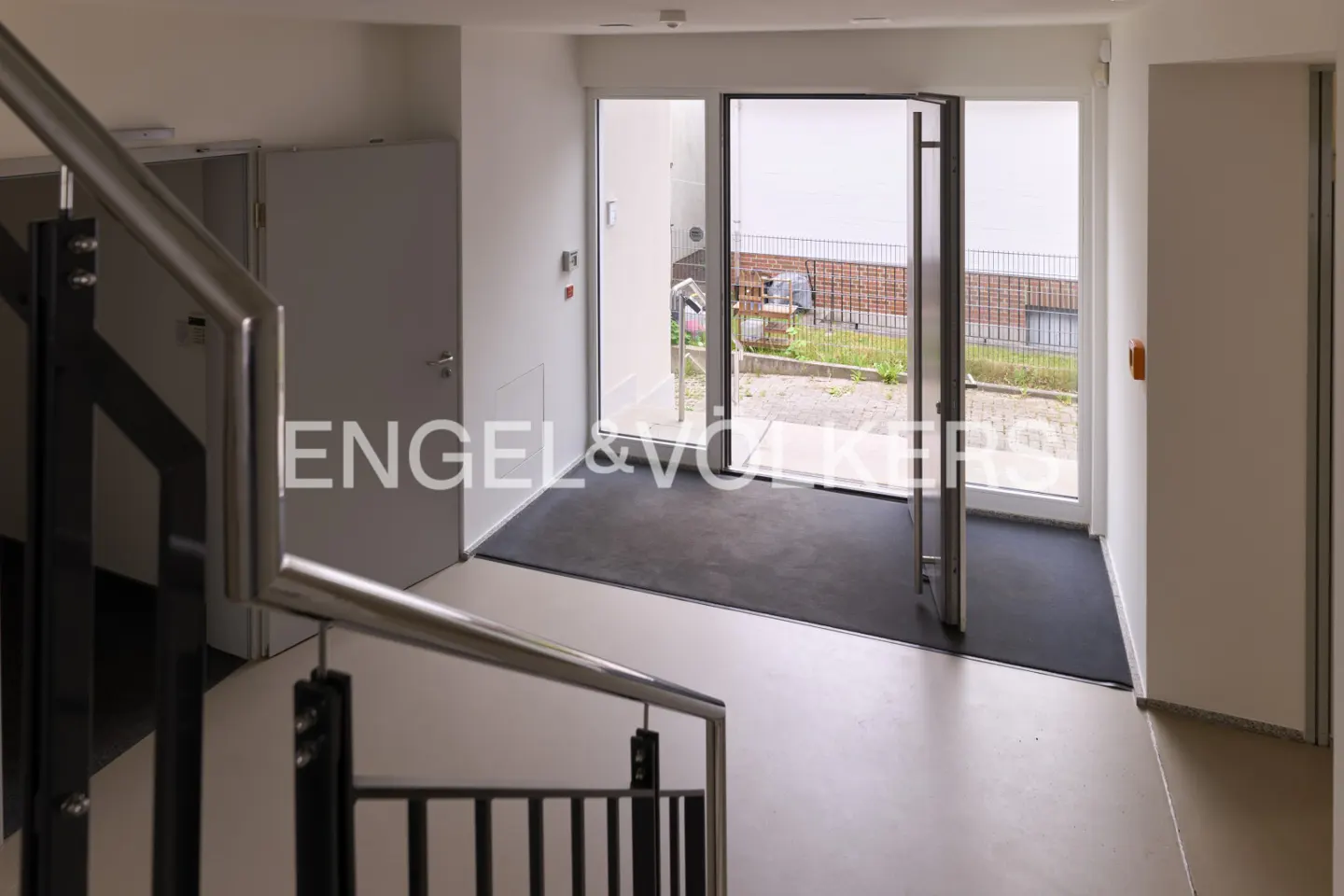 Interior view of a bright entryway with an open glass door leading outside. A staircase with metal railings is on the left. The floor is light beige.