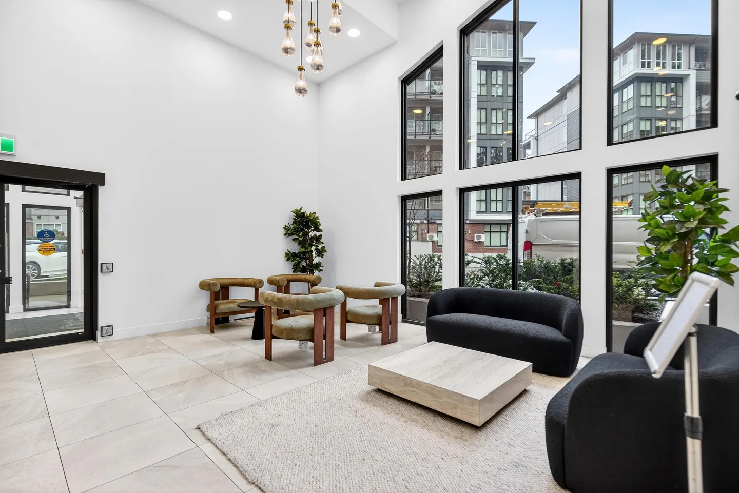 Modern lobby with white walls, tile floors, and black framed windows. Olive chairs, black sofa, and light wood table on a cream rug.