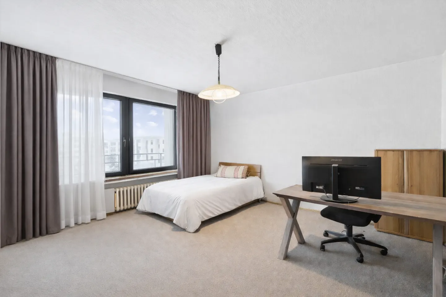 Bedroom with a single bed, desk with computer, and a window with gray and white curtains. A light fixture hangs from the white ceiling.