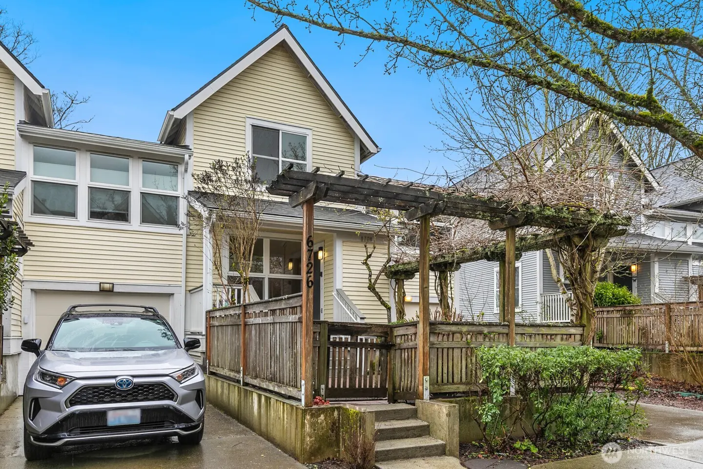 Exterior of a two-story yellow townhouse with a silver SUV parked in the driveway. A wooden pergola covers the front entrance.
