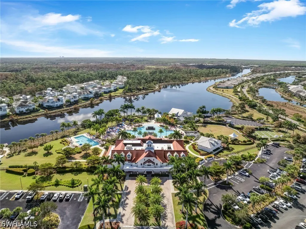Aerial view of a resort with a red-roofed building, pools, palm trees, parking lot, and a lake surrounded by trees under a blue sky.