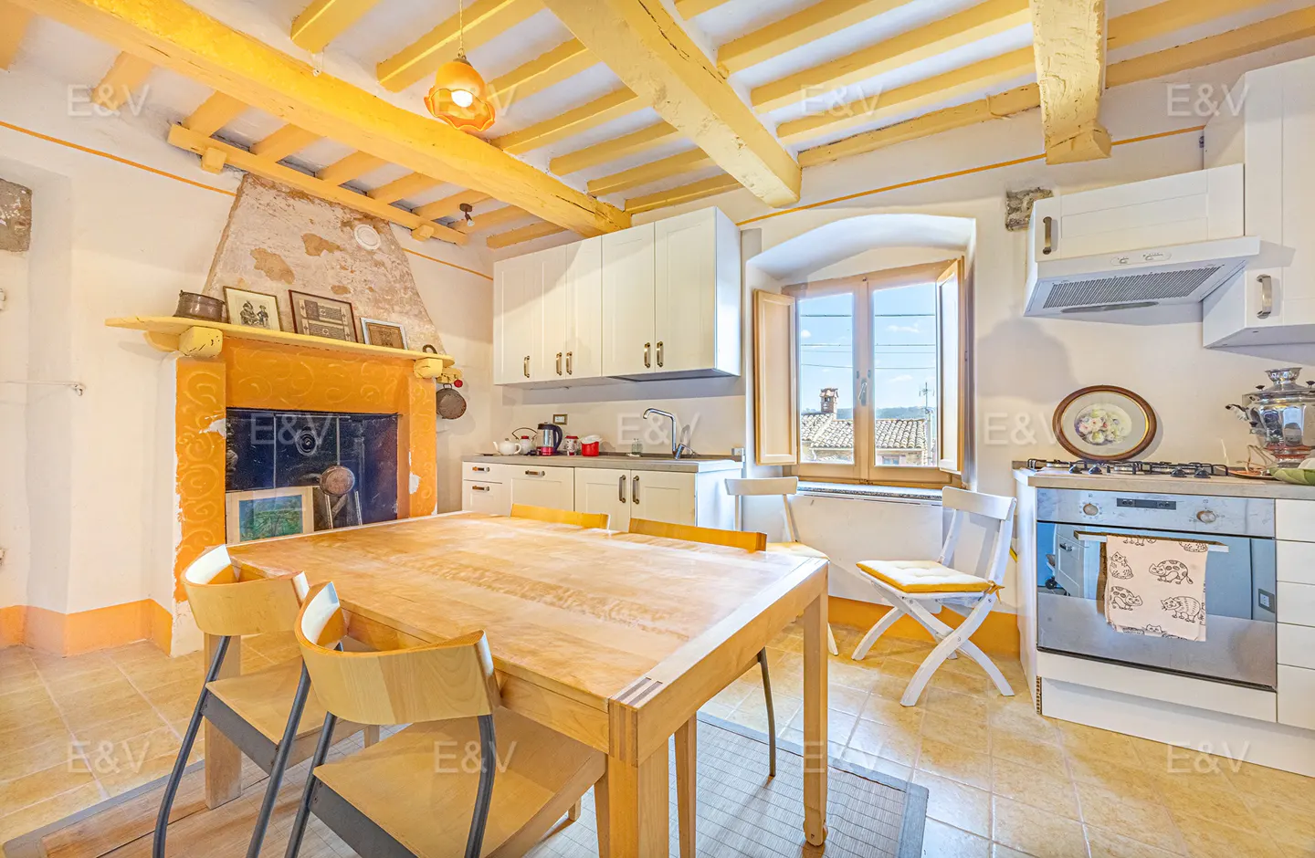 A bright kitchen with yellow ceiling beams, a wooden table with chairs, and white cabinets. A window overlooks a scenic view.