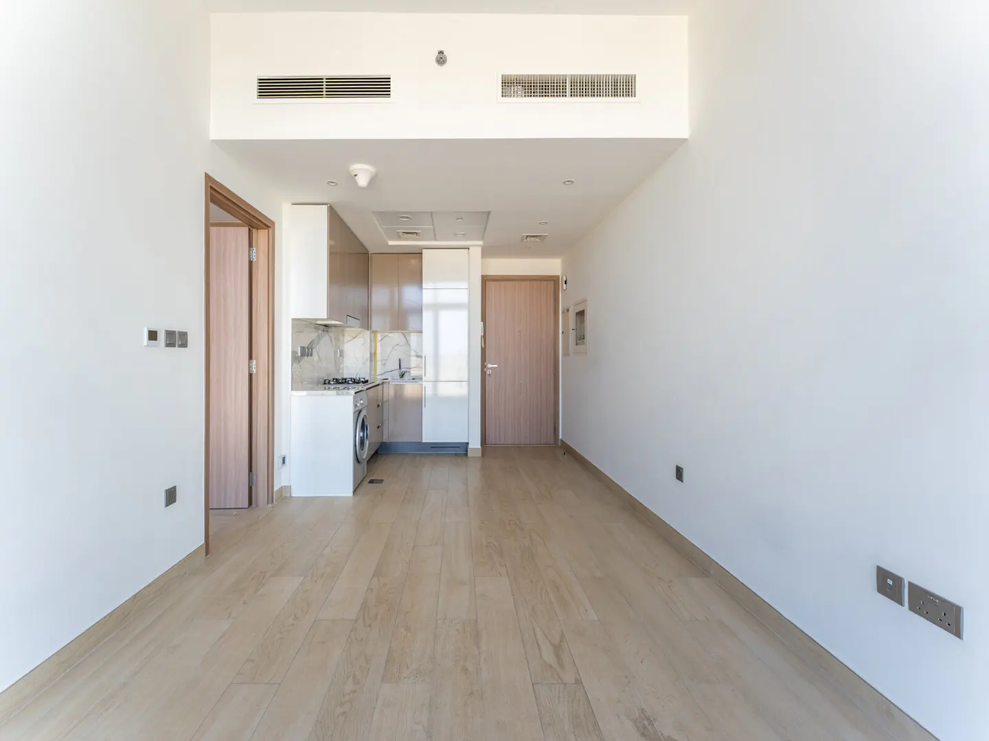 Bright, empty apartment with light wood floors, white walls, and a compact kitchen with a washing machine.