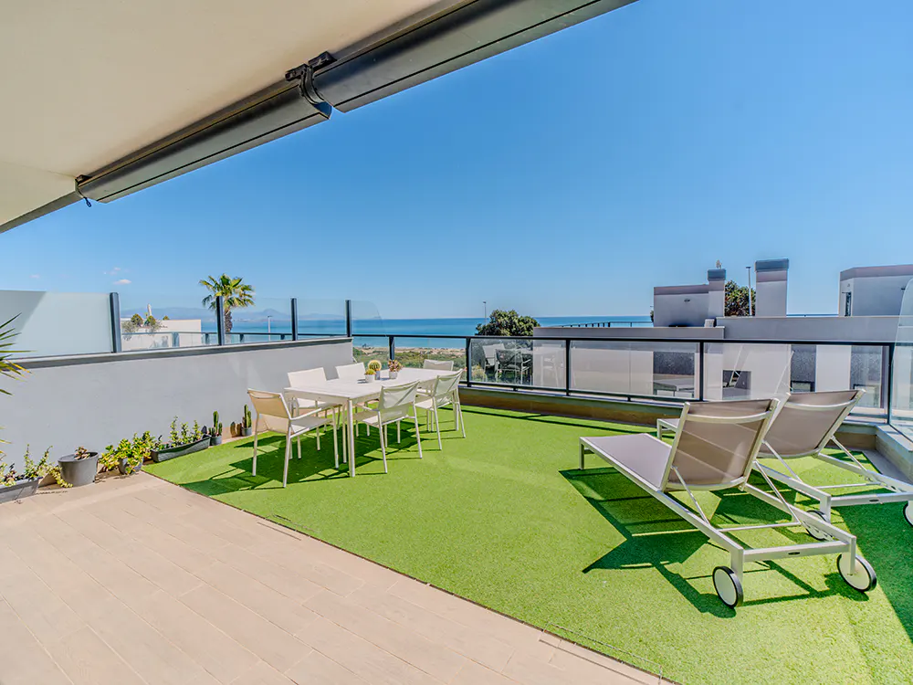 Outdoor patio with artificial grass, a white dining set, and two lounge chairs overlooking the ocean on a sunny day.