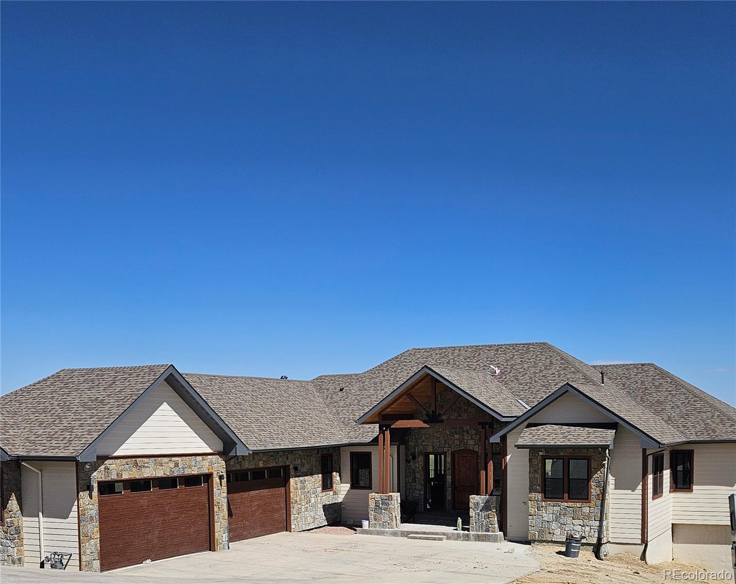 A single-story home with a stone and siding exterior, brown garage doors, and a gray shingled roof under a clear blue sky.