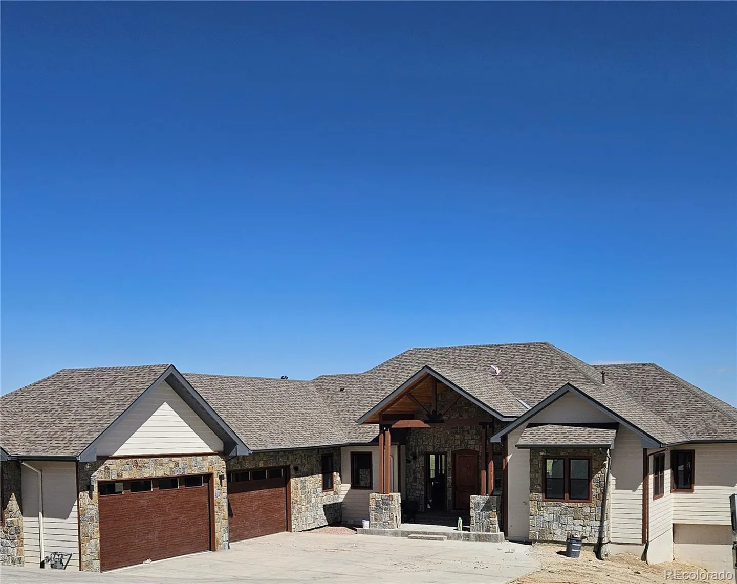 A single-story home with a stone and siding exterior, brown garage doors, and a gray shingled roof under a clear blue sky.