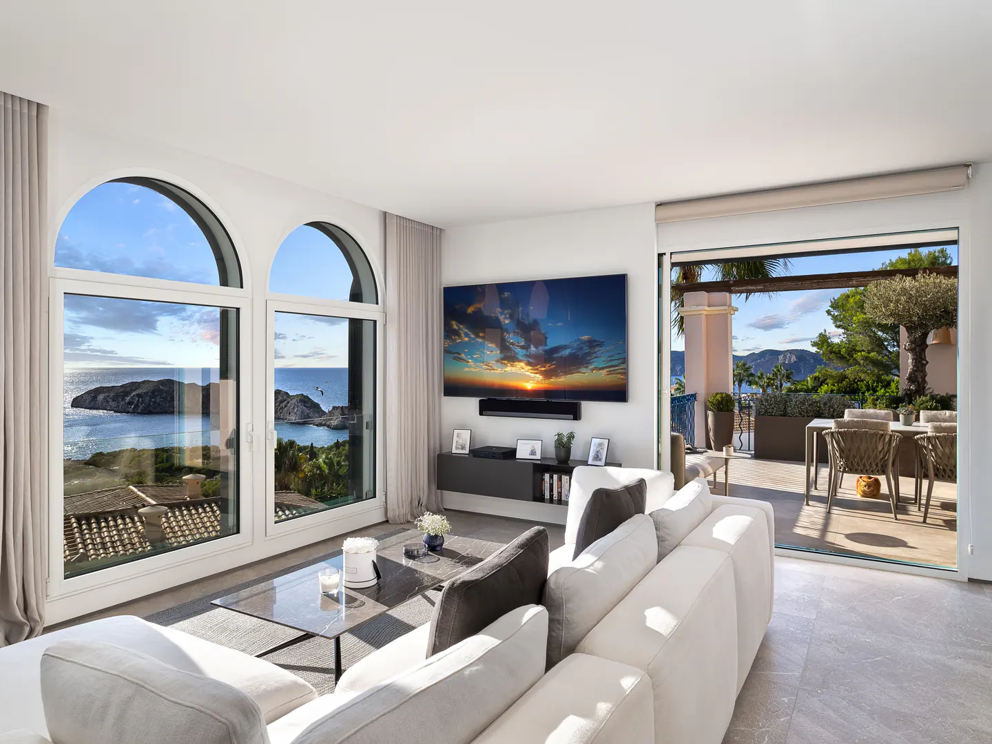 Bright living room with a white sofa, arched windows with ocean views, and a sliding door to a patio with outdoor dining.