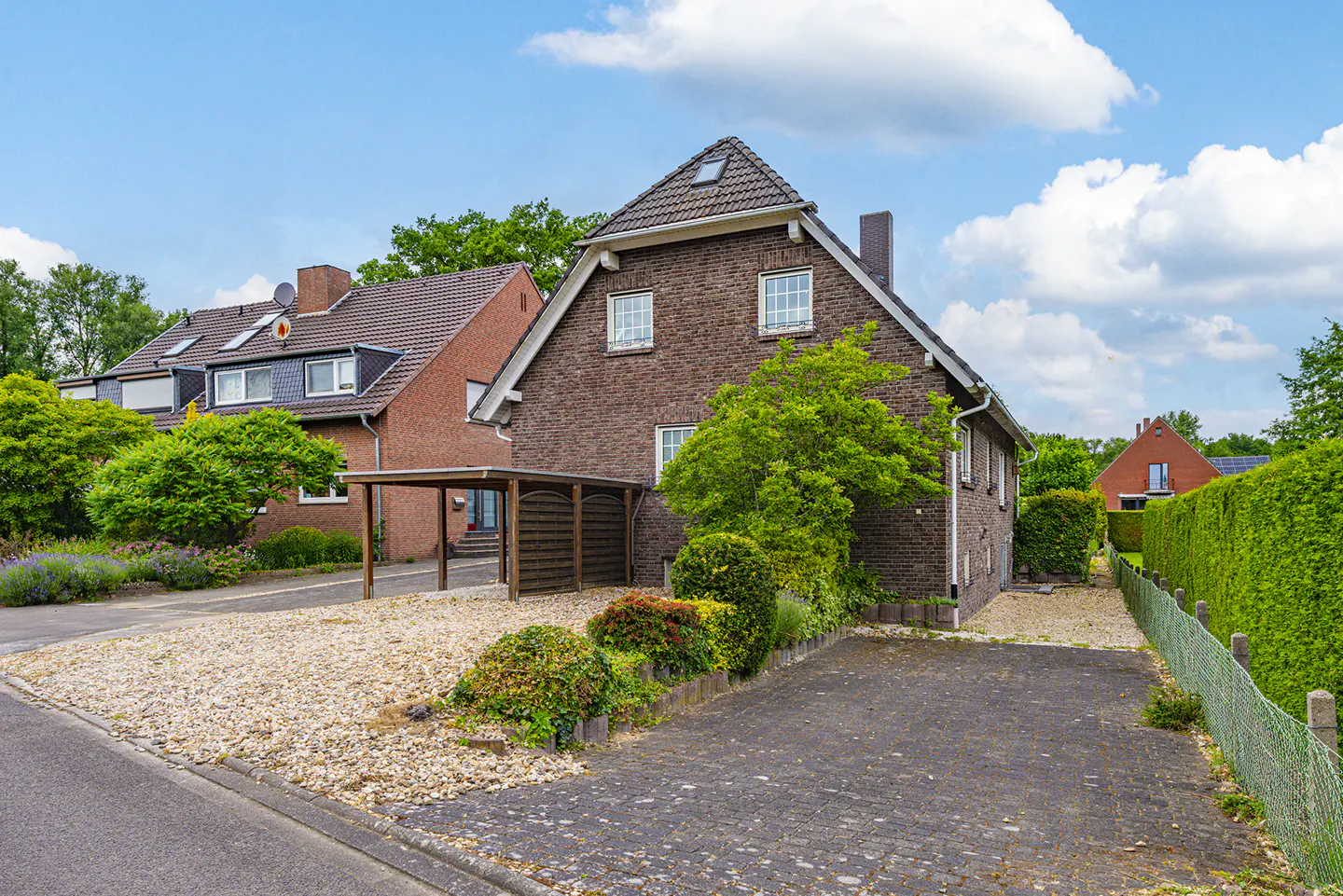A brown brick house with a carport, gravel driveway, and paved parking area under a bright blue sky with white clouds.