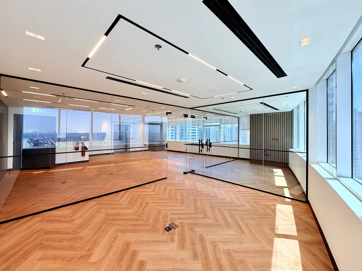 Empty office space with herringbone wood floors, glass walls, and black trim. Large windows offer a city view. Modern lighting fixtures on the white ceiling.