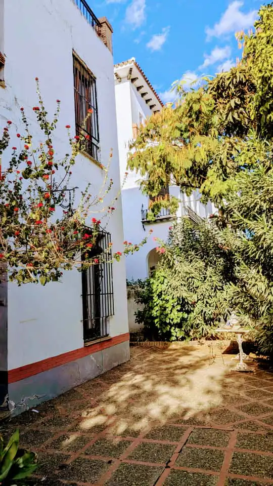 Exterior view of a white building with barred windows, a red-flowered bush, and a brick-patterned courtyard with a bench.