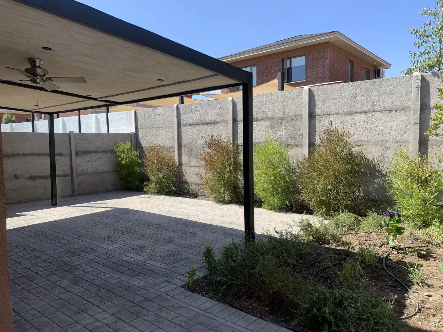Outdoor patio with a black metal pergola, ceiling fan, and gray brick flooring. A concrete wall with shrubs lines the back of the yard.