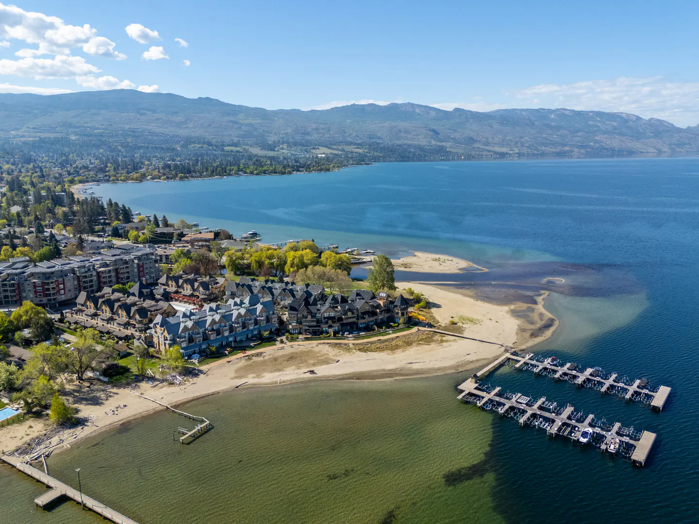 Aerial view of waterfront condos with a sandy beach, boat docks, and a large blue lake with mountains in the background.