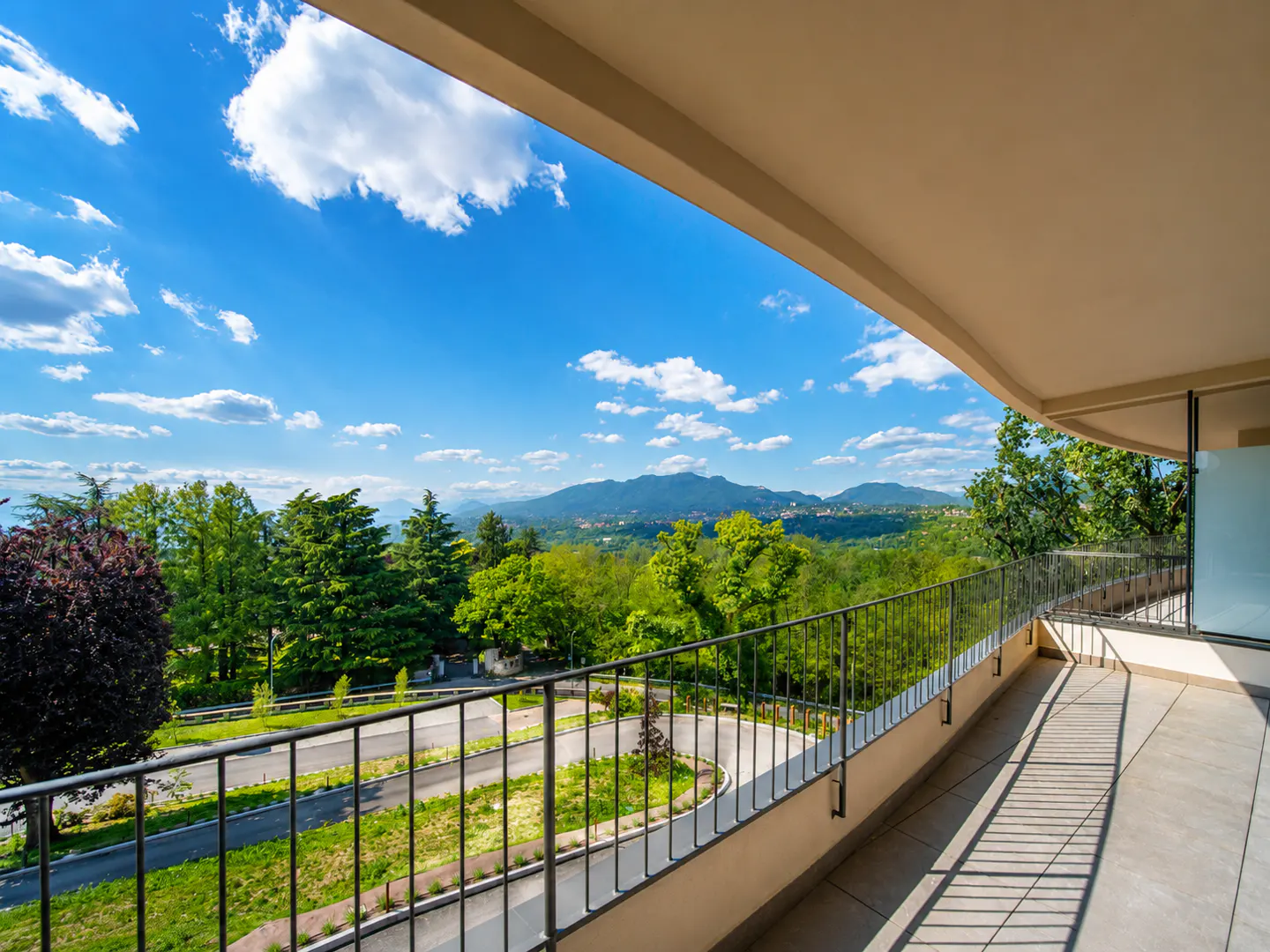 Balcony view with metal railing overlooking green trees, a road, and distant mountains under a blue sky with white clouds.