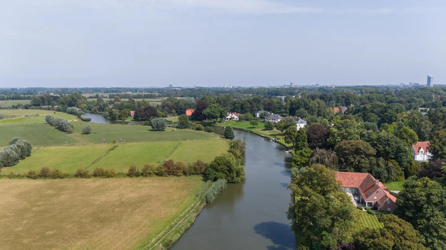 Aerial view of a river flowing through green fields and trees, with houses and a city skyline in the distance.