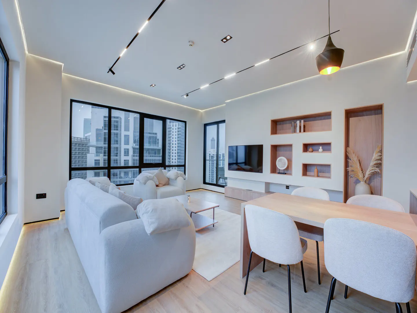 Bright, modern apartment interior with white sofa, dining table, and city views through large windows. Track lighting on the ceiling.