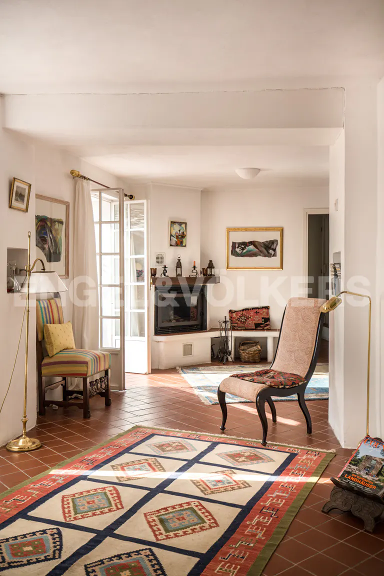 Bright living room with white walls, terracotta tile floor, and colorful rugs. A fireplace, chaise lounge, and striped chair add character.