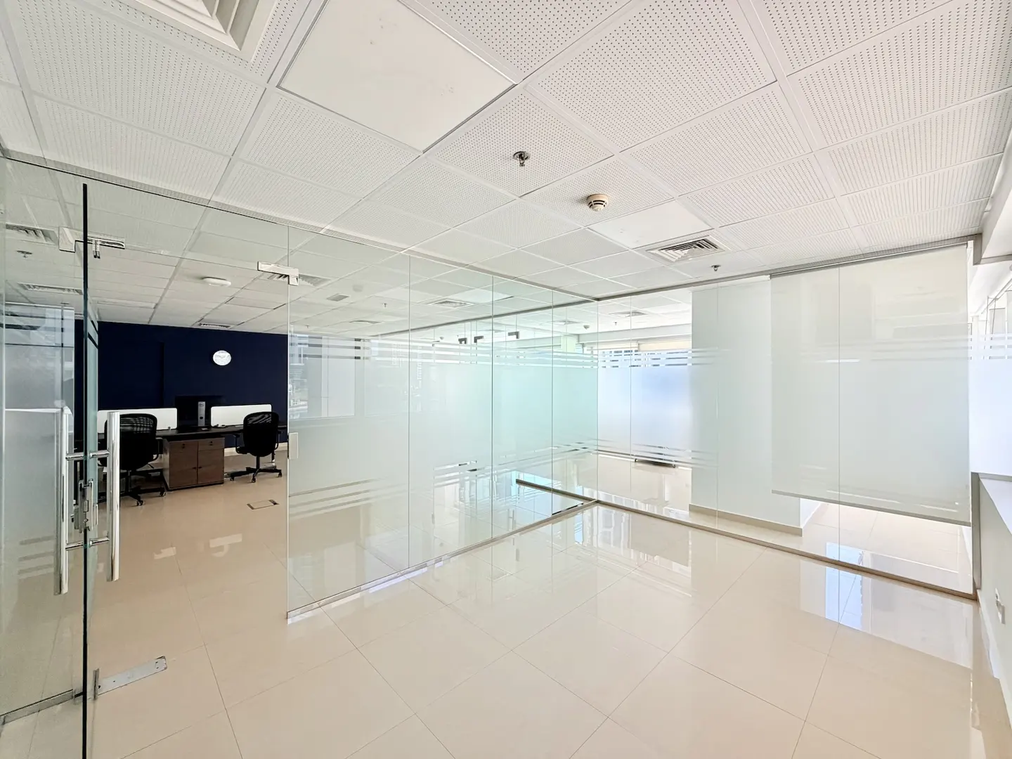 Empty office space with glass walls, beige tile floor, and white ceiling. Desks and chairs are visible in the background.