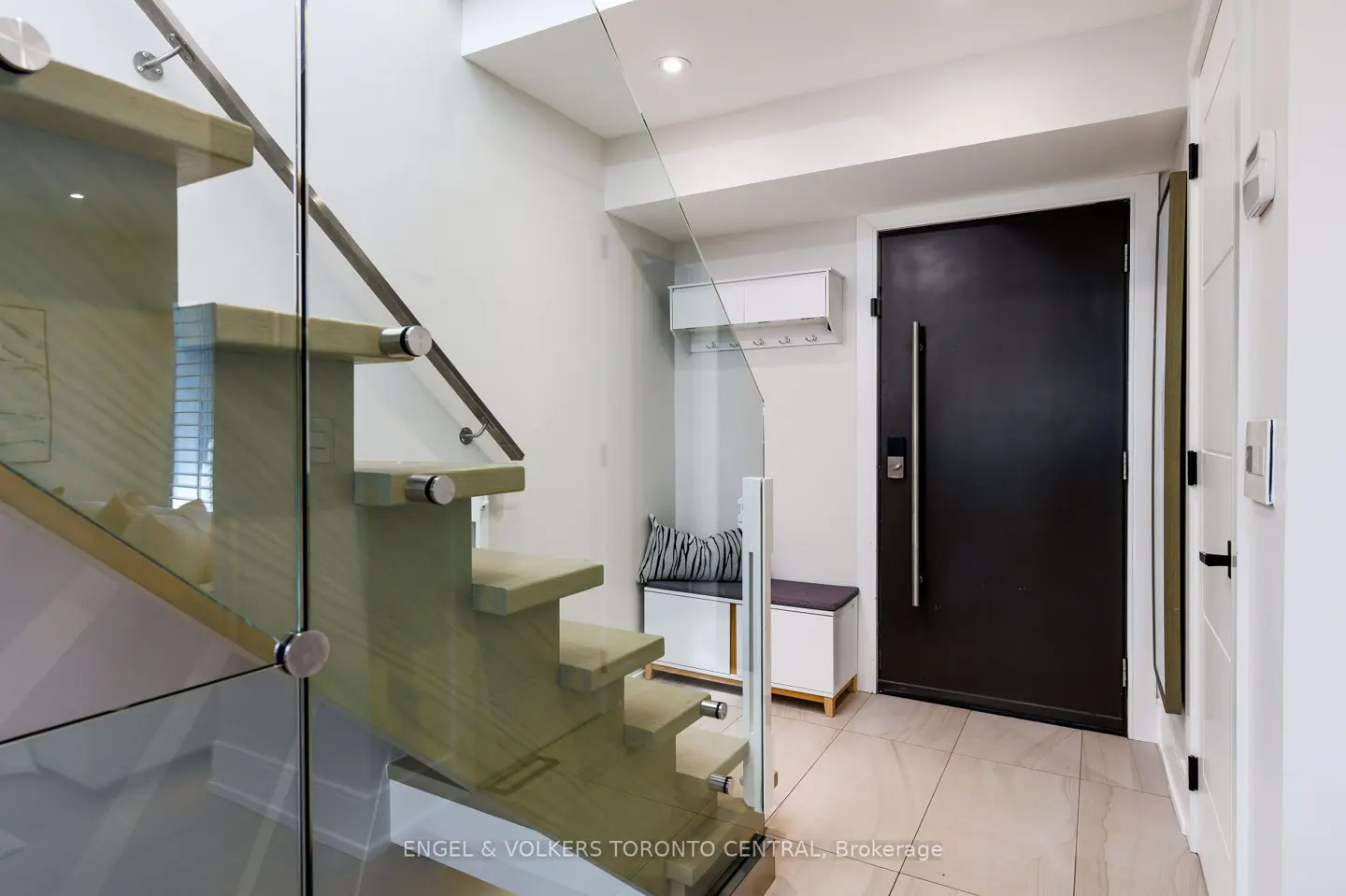 Modern foyer with floating stairs, glass railing, and black front door. A white bench and storage unit sit against the wall. Beige tile flooring.