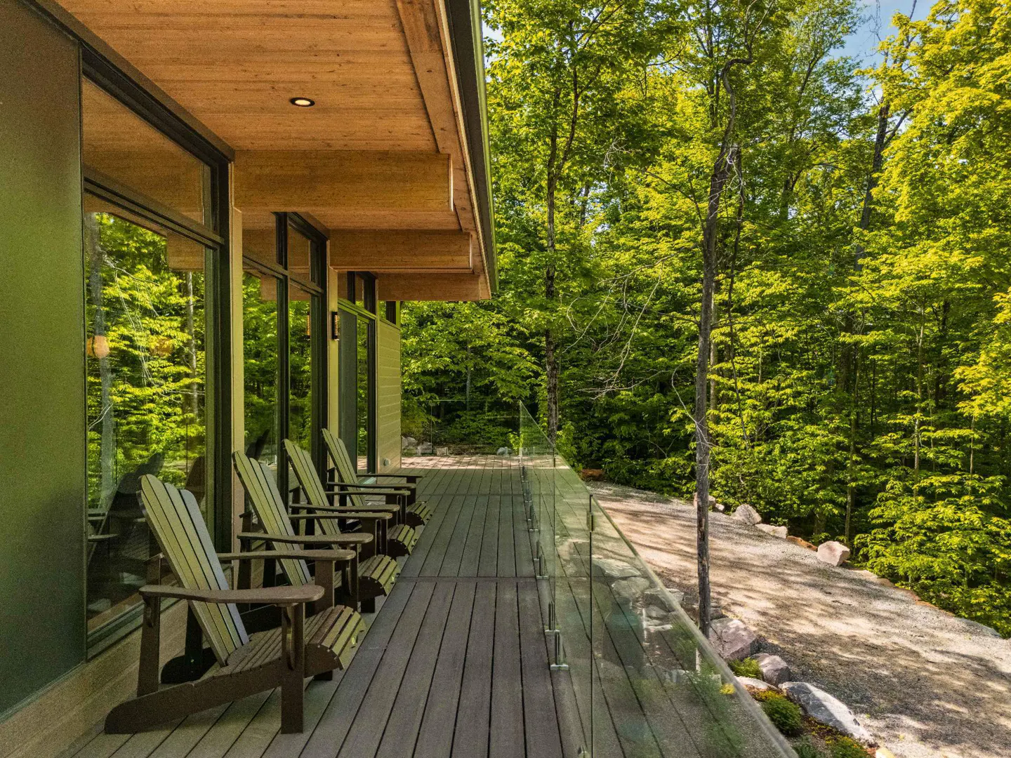 Modern wooden deck with Adirondack chairs overlooking a lush green forest and a gravel path.