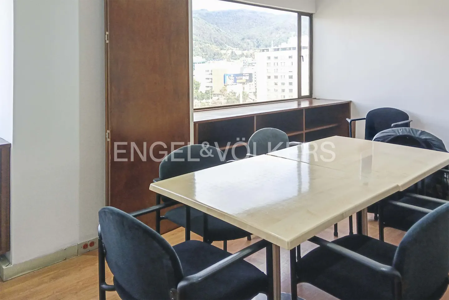 Office space with a large window overlooking a city. A table with black chairs is in the foreground.