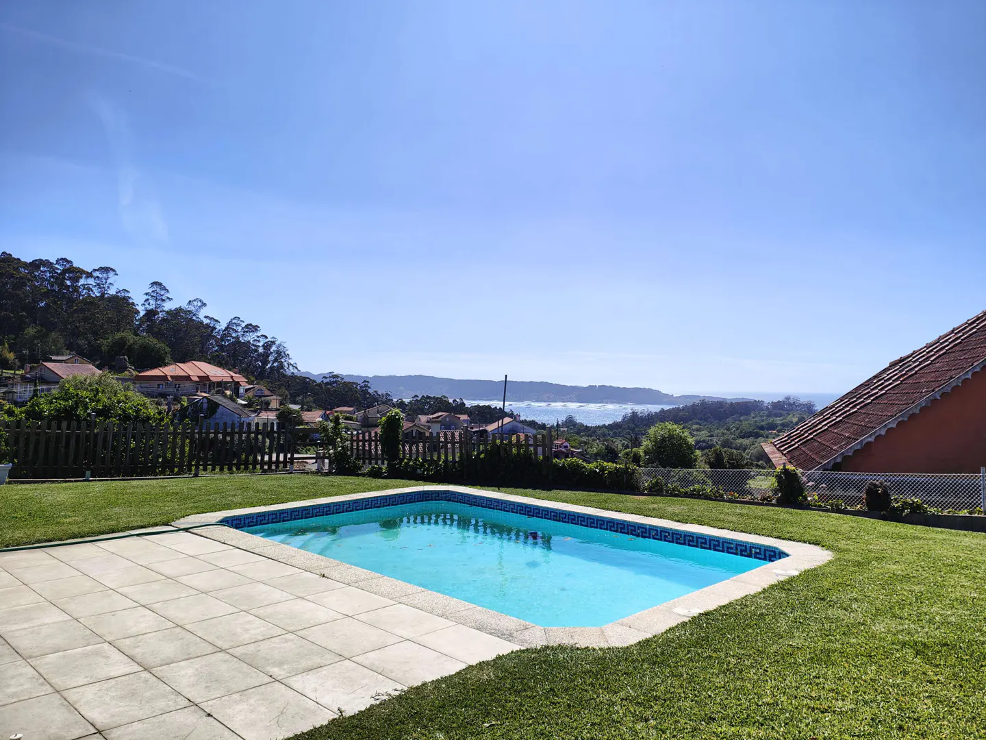 A backyard pool with blue water, surrounded by green grass and a tiled patio, overlooks a scenic view of houses and the ocean.