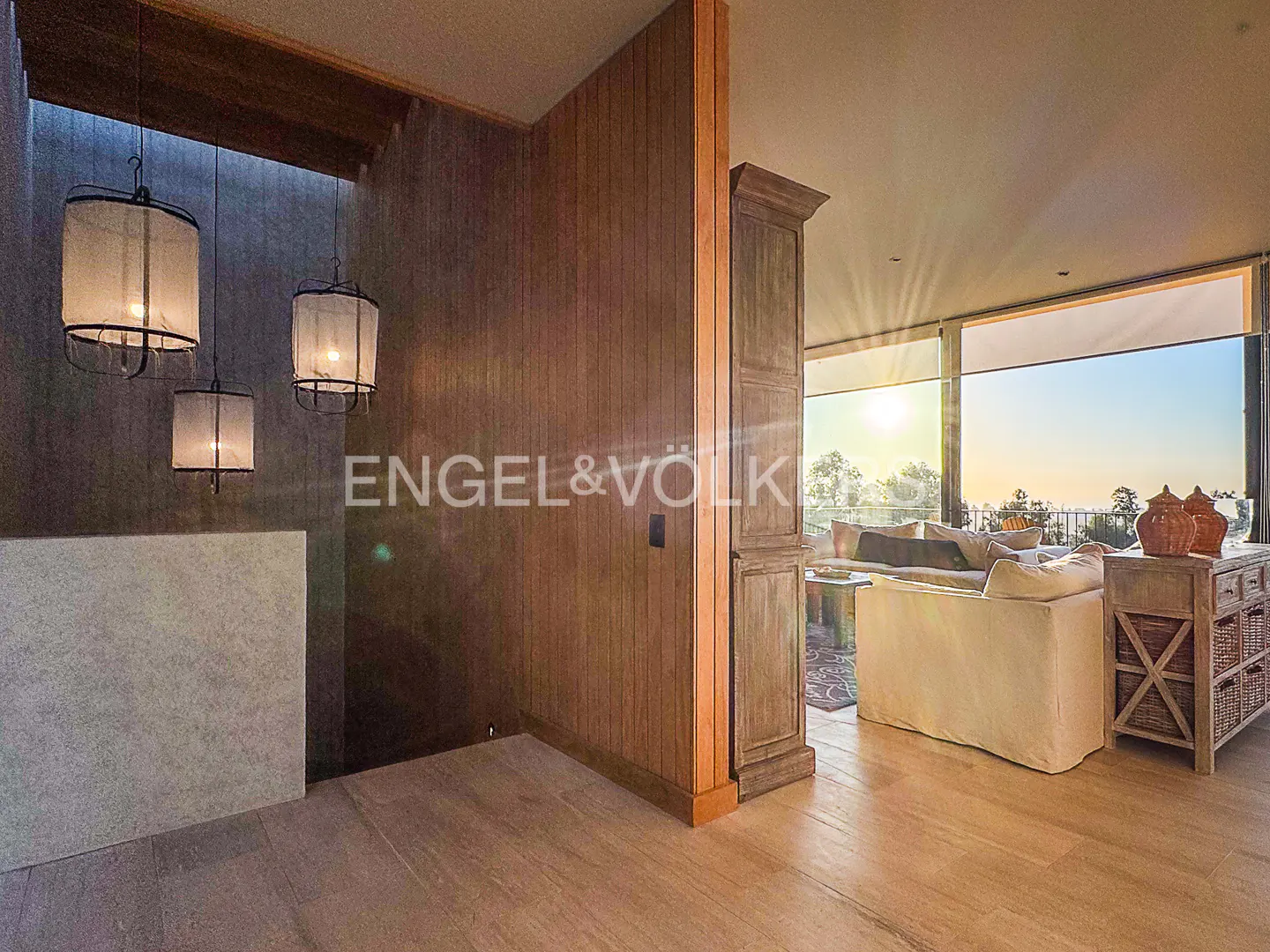 Interior view of a modern home with wood floors, walls, and hanging lamps. A living room with a white sofa and large windows is visible in the background.