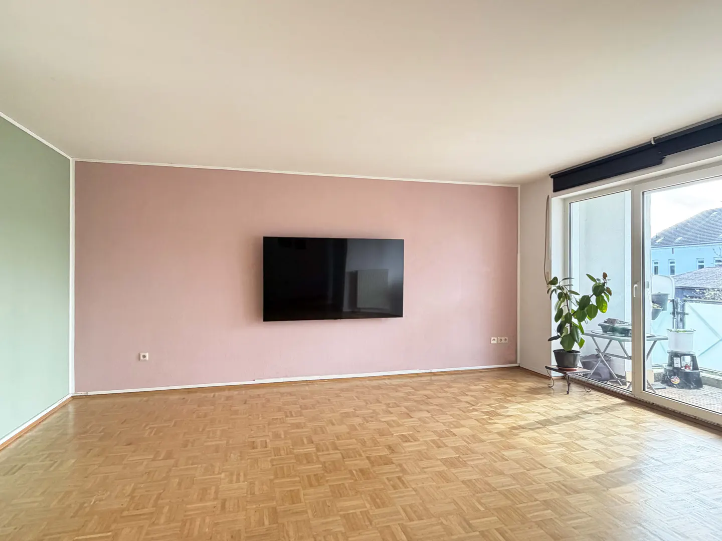 Empty living room with parquet floor, pink and green walls, a TV, and a balcony with a plant.